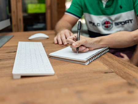 Keyboard and woman's hand holding pen writing in notebook while sitting next to a man at wooden desk to provide assistance with DVA Claims Brisbane - Contact VHC today