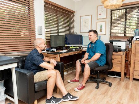 Male doctor sitting at wooden desk in medical rooms talking to male patient about providing World-class Veterans Health Care Help in Darwin - Contact VHC today