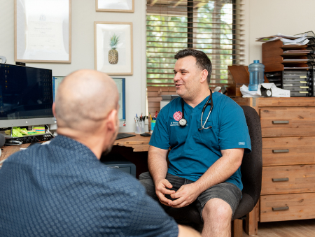 Male doctor sitting at wooden desk looking at computer and talking to male patient about veterans health care in Brisbane clinic - Contact VHC today