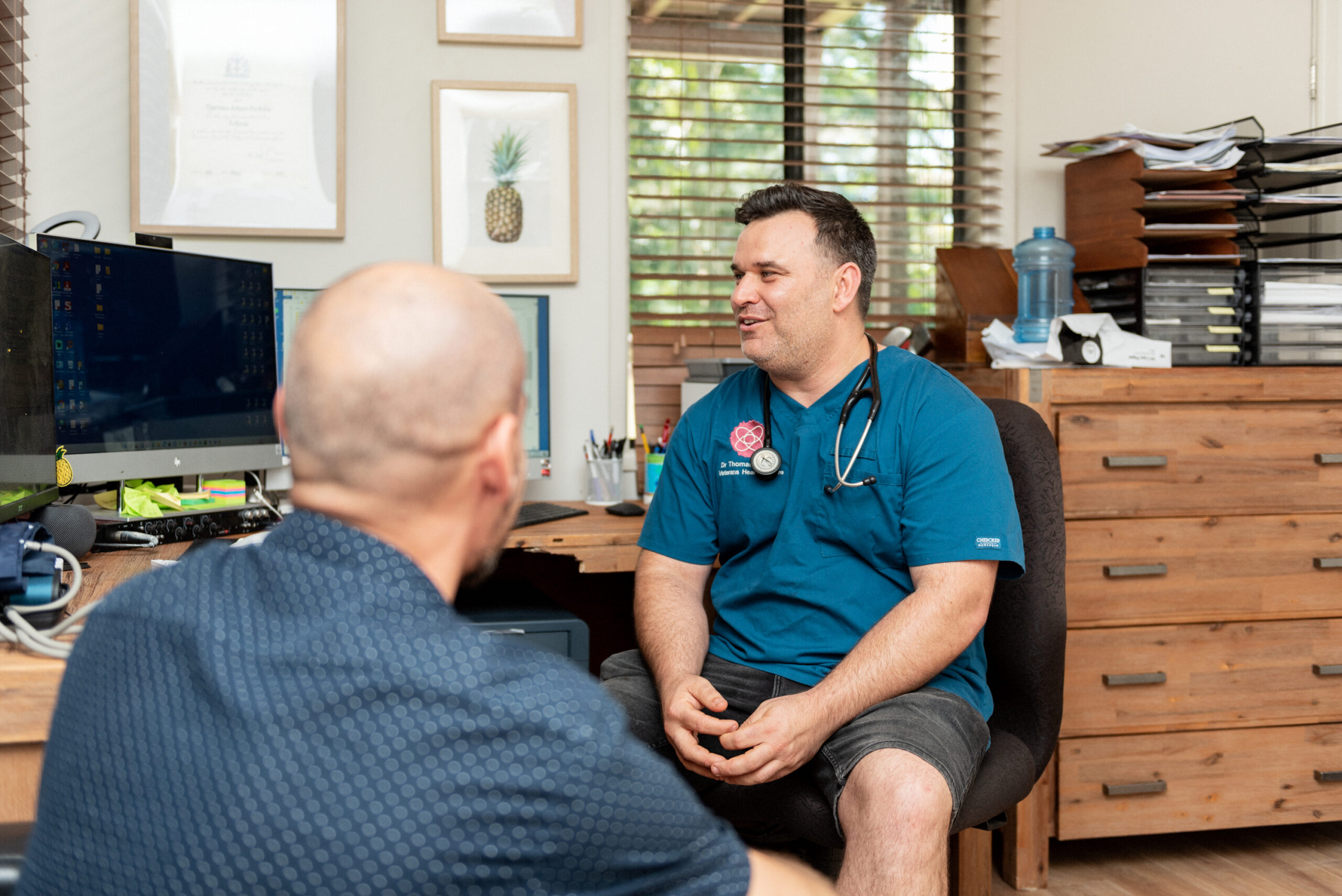Male doctor sitting at wooden desk looking at computer and talking to male patient about veterans health care in Sydney clinic - Contact VHC today